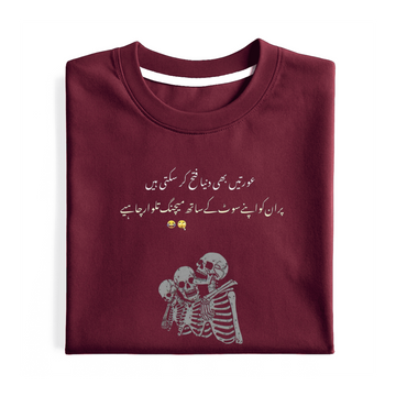 A folded maroon crewneck t-shirt with white Urdu text and a white skeleton graphic on the front, displayed on a clean white background.