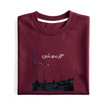 A folded maroon crewneck t-shirt with a graphic print on the front. The print features white Urdu script and a small illustration of a street scene at night with streetlights and cars.